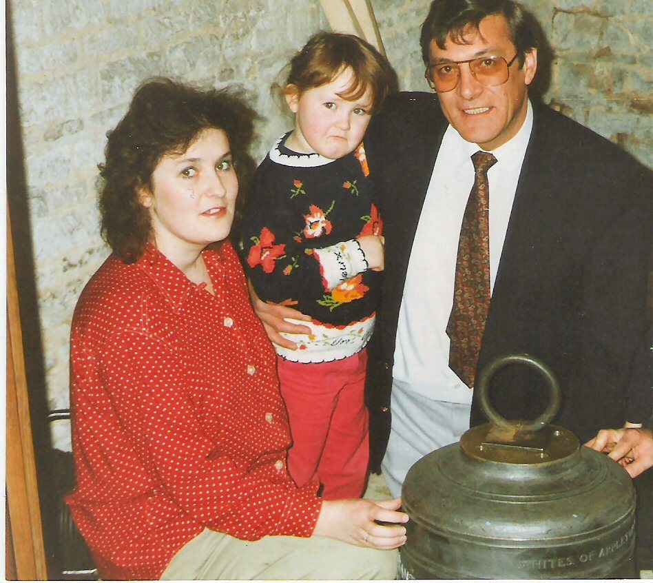 A family with a young child beside a new church bell standing on the floor, its rim marked Whites of Appleton
