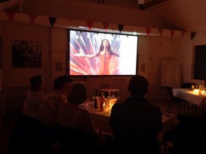 A film night in the hall, diners at candlelit tables watching a big screen
