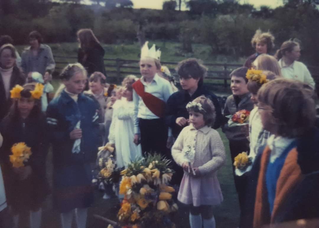 Children in flower garlands with posies of daffodils, gathered around the May King in a paper crown and red sash, with the May garland in front