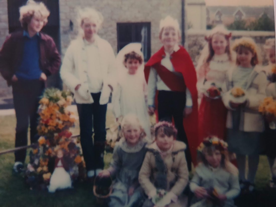 A group of children around the May King and Queen, beside a tall May garland of spring flowers with a doll at its foot
