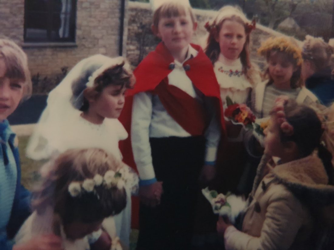The May King in a red cape and the May Queen in a white veil, with children wearing flower garlands and carrying posies