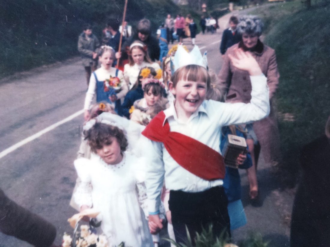 Children in a May Day procession along a village road, led by a waving boy in a paper crown and red sash holding hands with a girl in a white veil