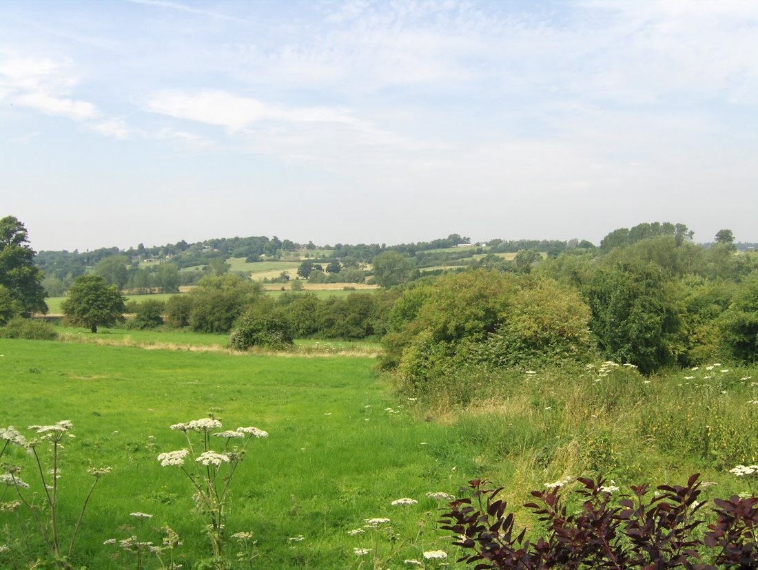 Green fields falling away to hedges and trees, with the far side of the valley rising beyond